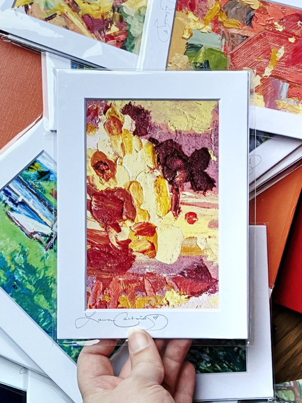 Top down view of a hand holding up a colorful matted photo art print over a jumbled pile of other matted prints.